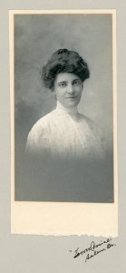 A photograph portrait of a woman from the shoulders up wearing early 1900s dress, facing the camera and smiling slightly.