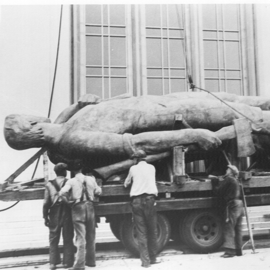 a black-and-white historical photo of the Oregon Pioneer (AKA Gold Man) laying horizontally on a truck bed and a group of men in 1930s clothing looking at the statue.